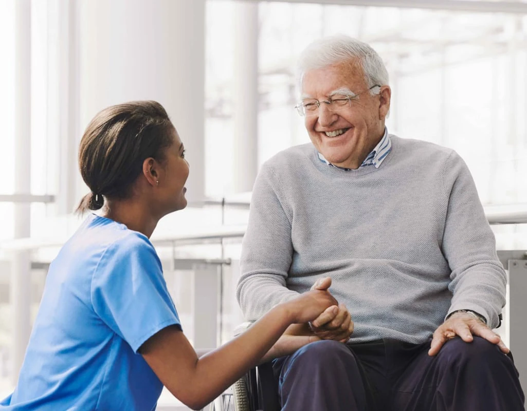 A female nurse kneeling next to an elderly man in a wheelchair and they are having a conversation.