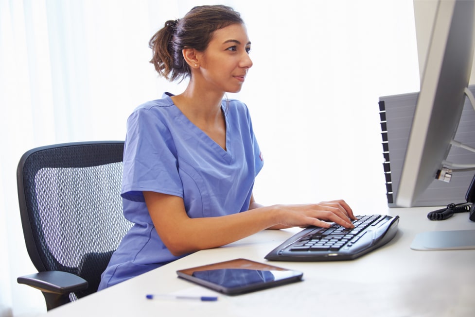 A woman in blue scrubs is alone typing on a keyboard in front of a keyboard