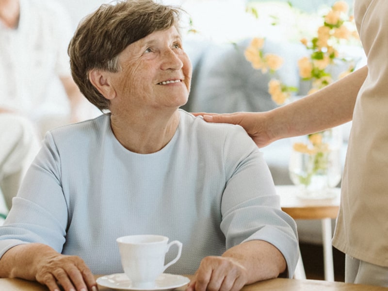 In the foreground an elderly woman is having a cup of coffee