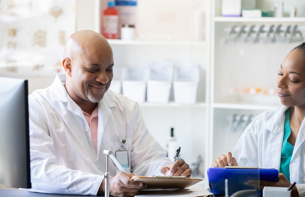 Two doctors, one male and one female, are seated having a discussion, while the male doctor is writing on a clipboard