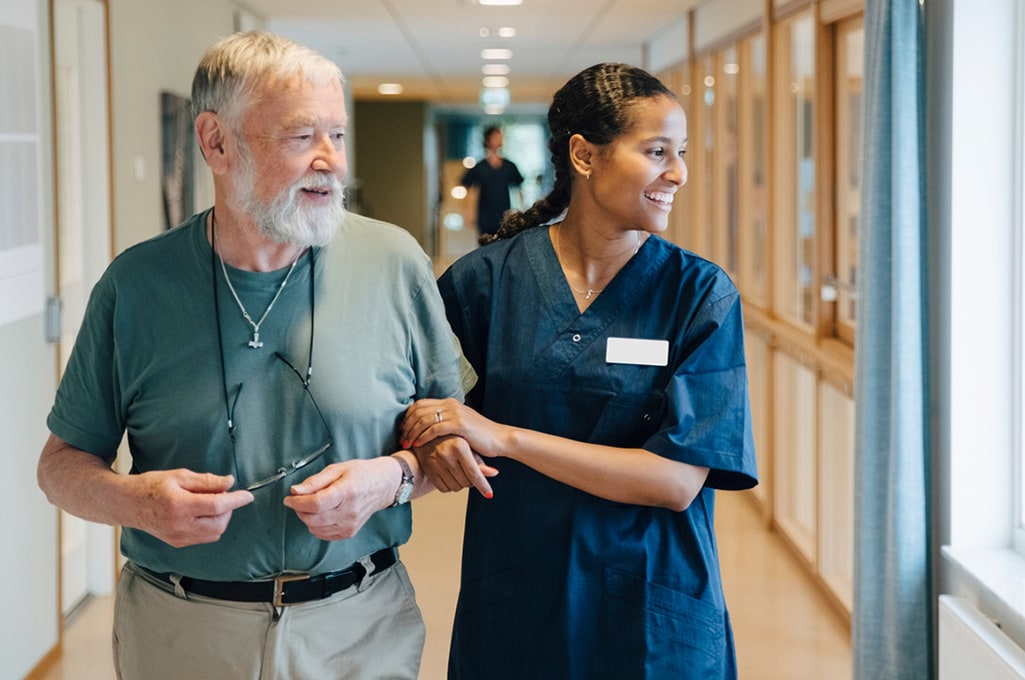 A female healthcare worker is assisting an elderly man who is walking