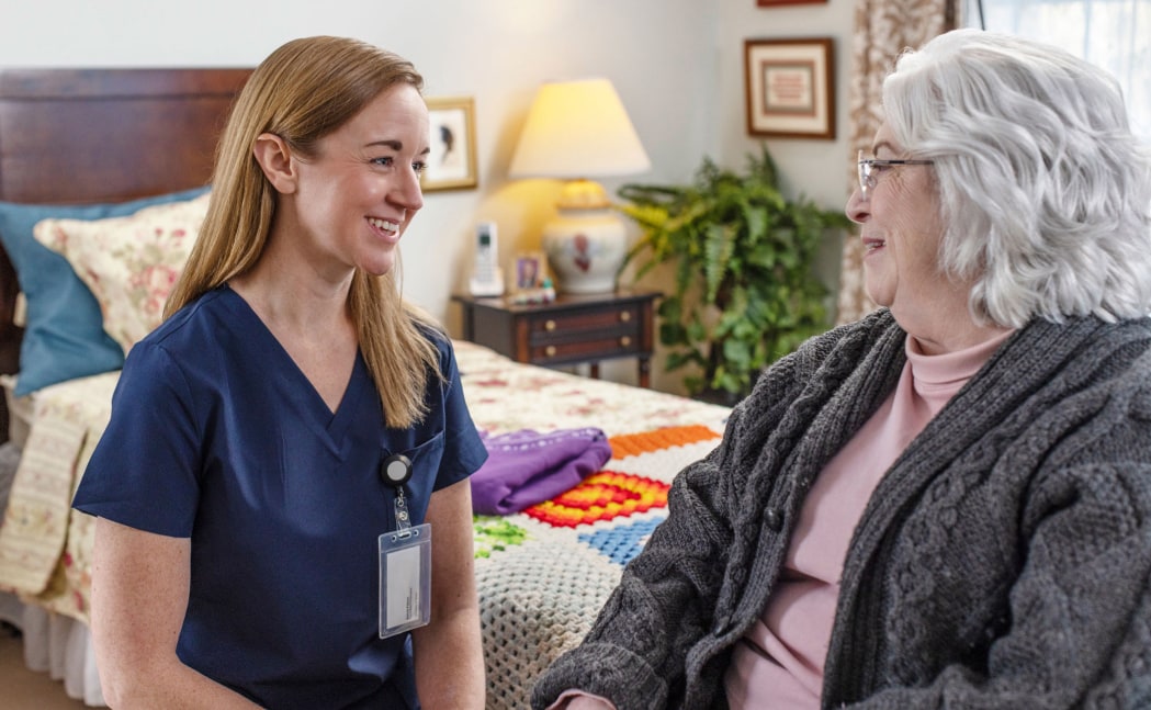 A female health care worker is having a seated conversation with an elderly woman in a bedroom