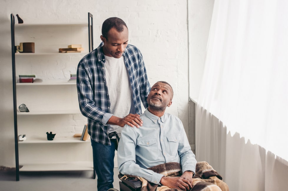 A younger man is looking over an older man who is seated in a wheelchair