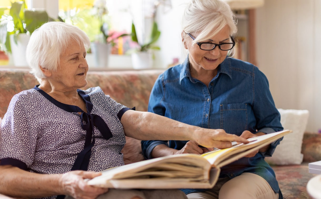 Two elderly women sitting on a couch, engrossed in a book, one of them sharing it with a younger loved one.