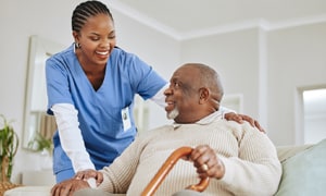 A man with a cane sitting in a chair is being helped by a nurse.